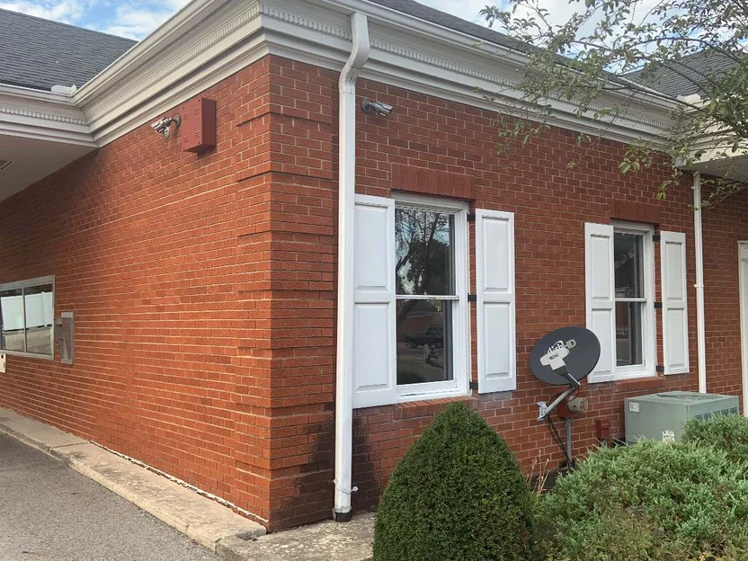 The bank in 2019 seen from the drive, two shuttered windows and a white downspout on red brick under a white cornice, a satellite dish fixed to the wall
