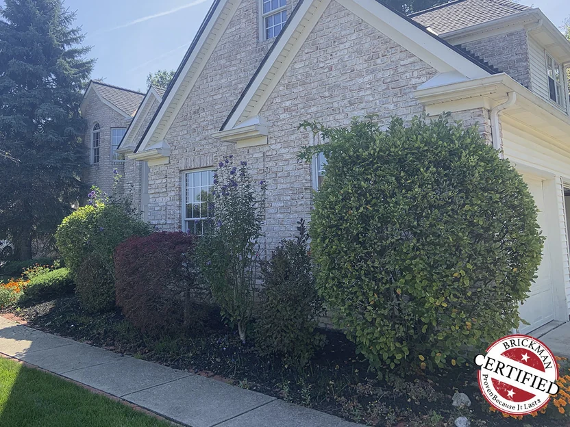 The gable end of the house in 2019, the brick an even cream and tan range from the flower bed up to the eaves, with the certification stamp in the lower corner