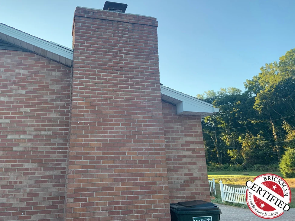The chimney from the driveway in 2019 with the house wall running away beside it, a Brickman Certified stamp printed over the lower right of the frame