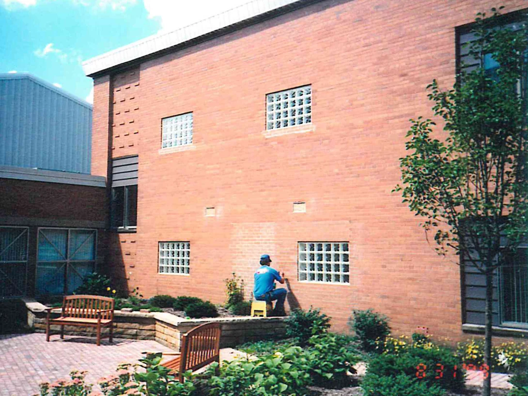 An applicator working the courtyard elevation from a low stool in 1998, the two-story brick wing rising above a paved garden
