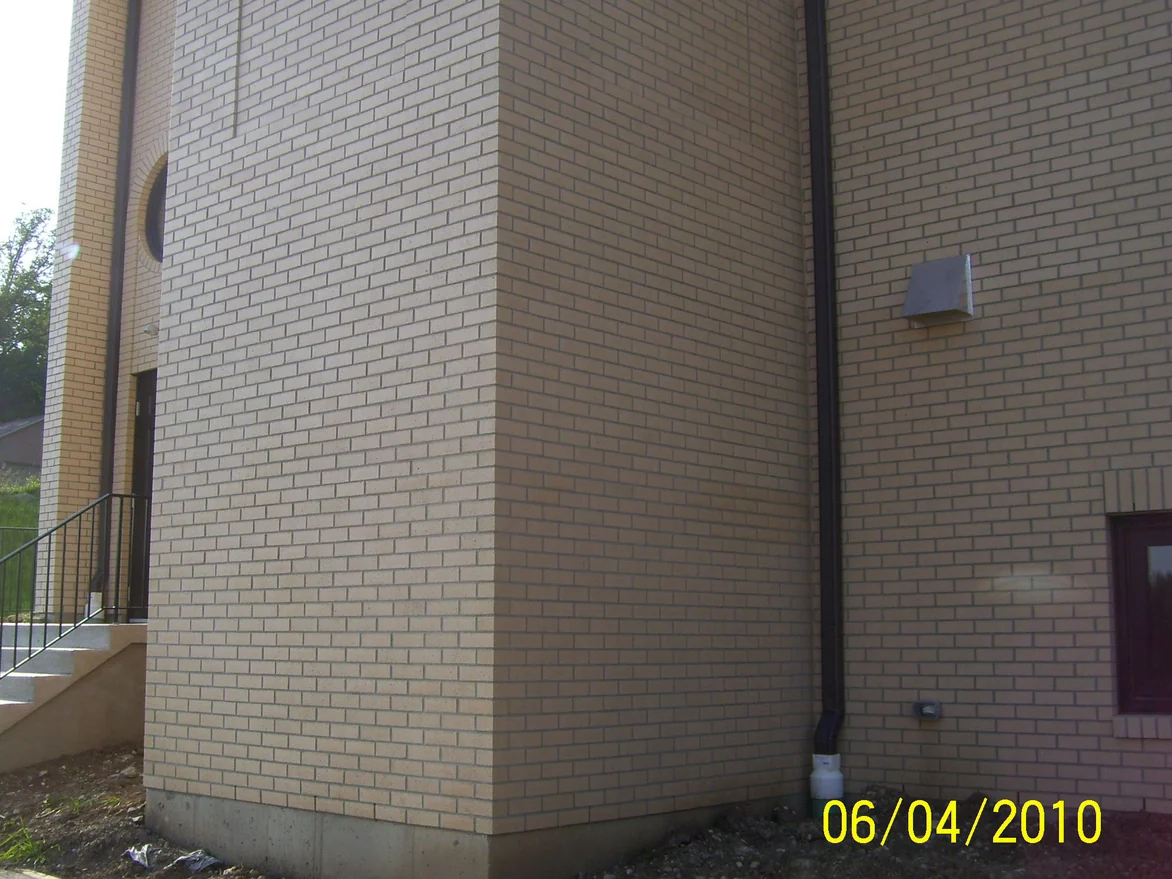 An outside corner of the church in 2010, warm tan brick above a concrete base with bare graded ground at the footing