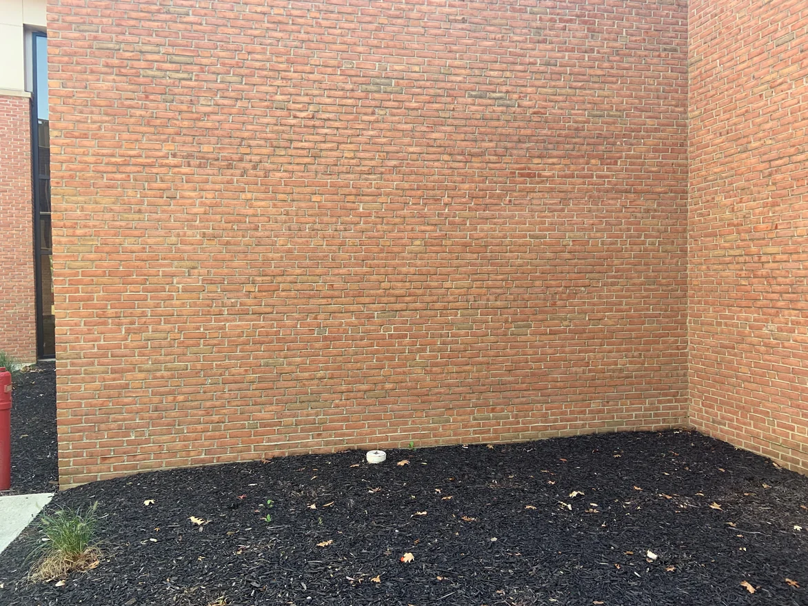 A long blank stretch of the brick wall meeting a return wall above a mulch bed, every course reading in the same red range