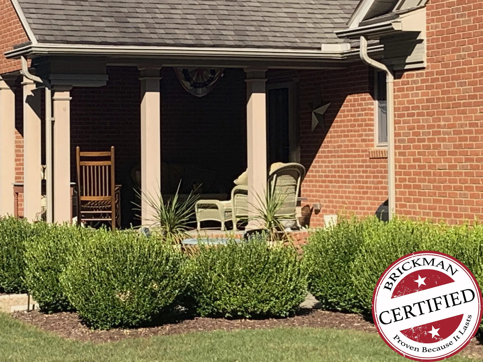 The covered porch in 2019, grown boxwood along its front and the brick wall beside it reading as one even red range, with a Brickman Certified stamp printed over the lower right of the frame