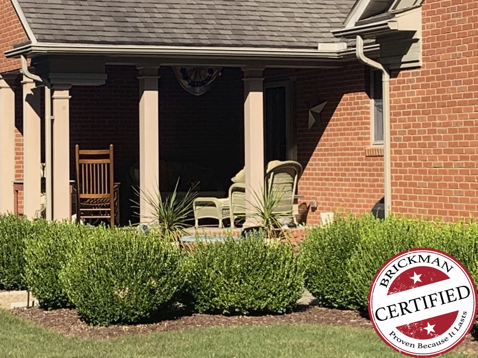 The covered porch of the house in 2019, grown boxwood along its front and the brick wall beside it reading as one even red range, with the certification stamp at the lower right of the frame.