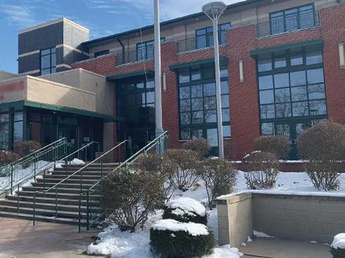 The front of the community center in 2021 under snow, a long red brick elevation with tall green framed windows, a flagpole and steps with green railings up to the entrance