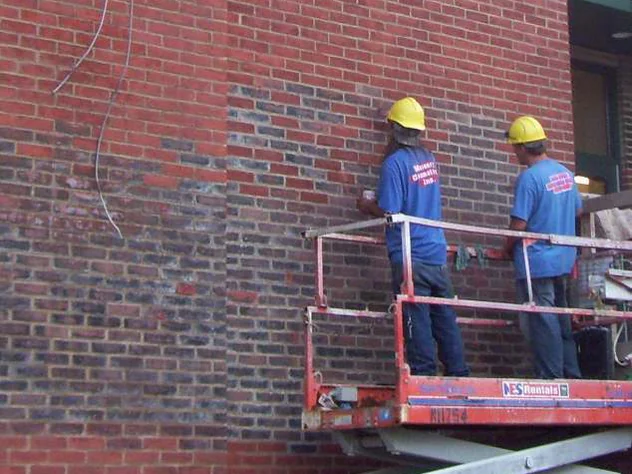 Two applicators in hard hats on a scissor lift in 2011 brushing stain onto a wall, the courses above and left of them still dark and the courses they have finished already red