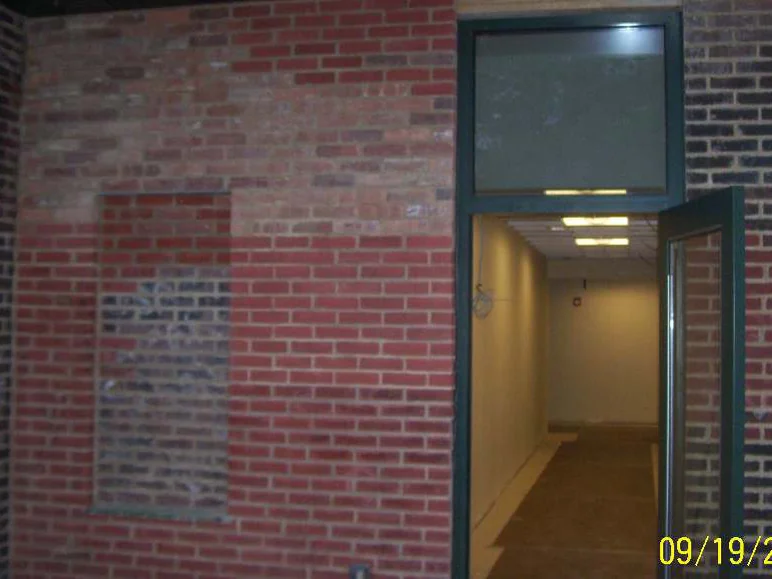 A recessed vestibule in 2011 with a green framed door propped open, the new brick around it dark and patchy against the older red brick and a grey block wall to the right, camera dated 09/19/2011