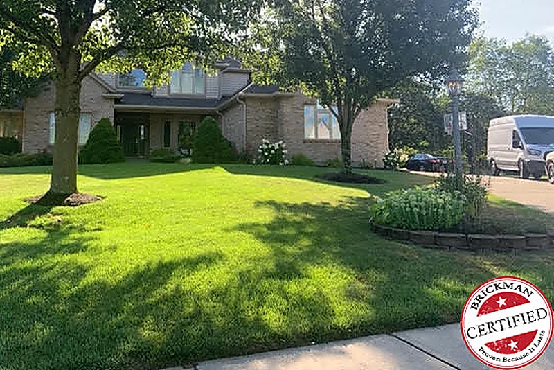 The house from across its lawn in 2019, grown trees over a mown yard and the brick behind them still reading as a range of light and dark units, with the certification stamp at the corner of the frame