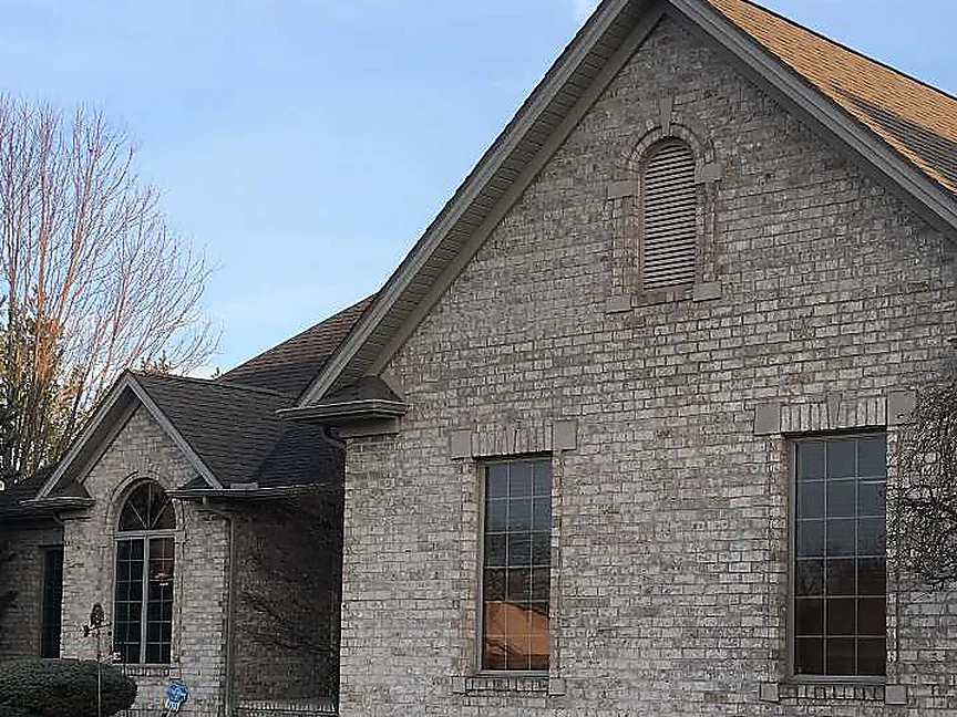 The house in 2019, buff brick running from the arched louver in the gable peak down past two tall windows to the lawn and the front walk