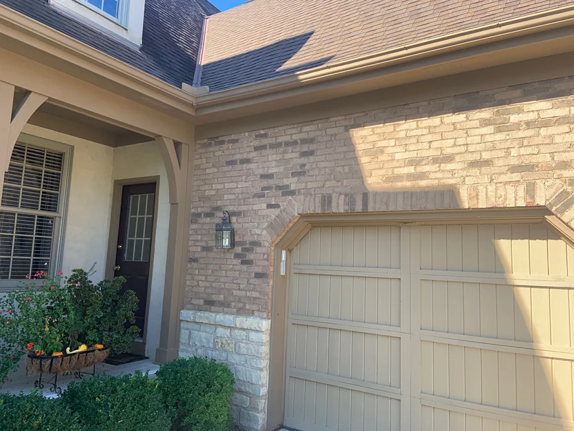 The entry in 2019, a dark front door under the stucco porch with the stained brick of the garage wing running away to the right above a stone base