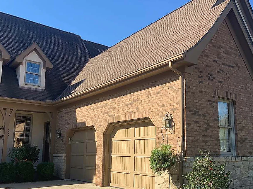 The same front of the house in 2019, brown brick from the dormered roof across the gable to two arched garage heads, with shrubs and a planted hanging basket at the base