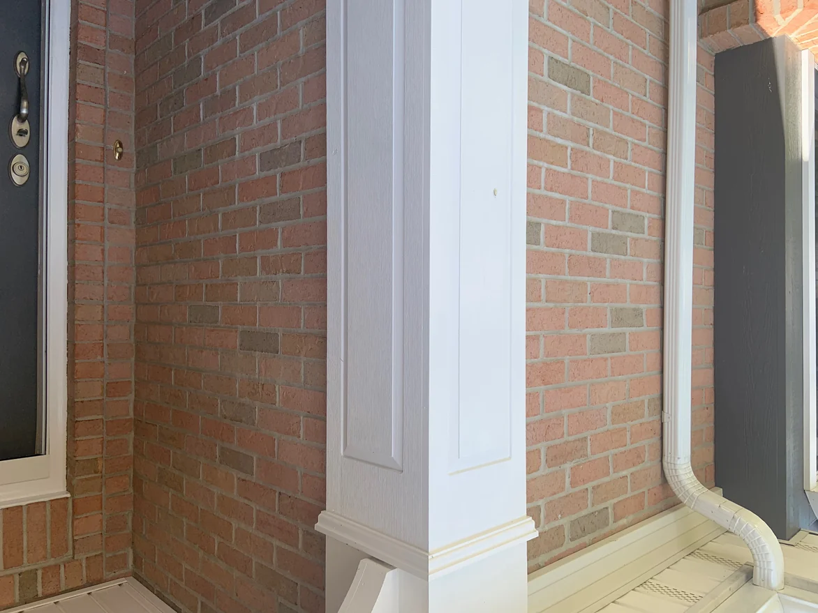 The brick beside the front door in 2019, an inside corner of the porch running back to a white column and a downspout, the units an even light range
