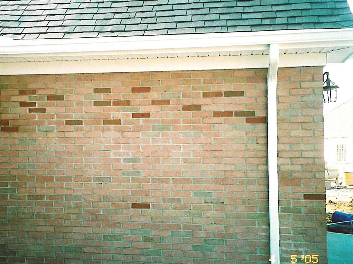 A gable end wall of light tan brick in 2005 with a scatter of much darker red units through the upper courses, a white downspout at the corner