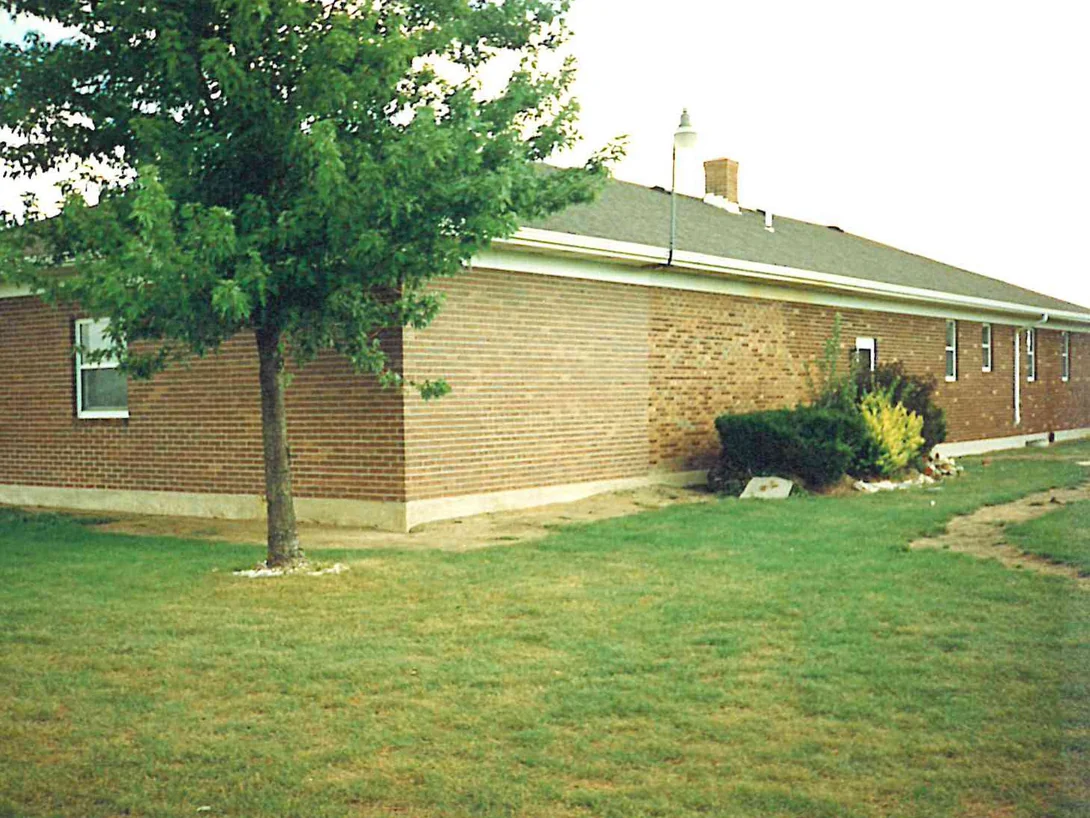The corner of the ranch house in 1995 under a young tree, the long wall beyond it showing a paler block of brick beside the shrubs where the addition meets the original