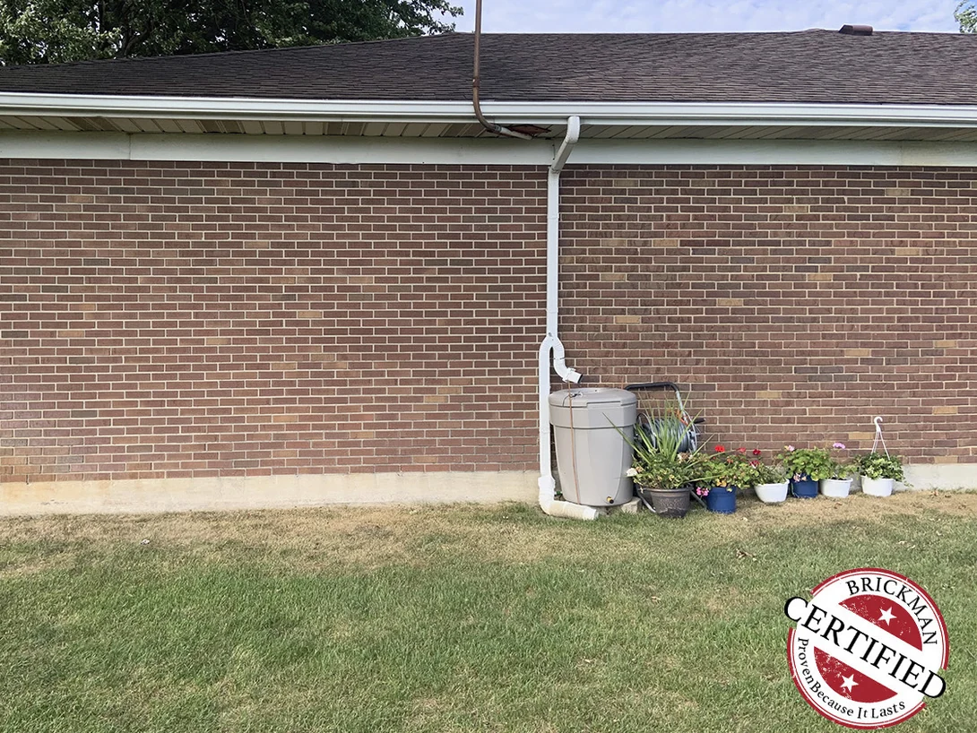 A blank run of red brown brick wall in 2019 under the eave, with a downspout, a rain barrel and potted flowers at its foot and the certification stamp in the corner