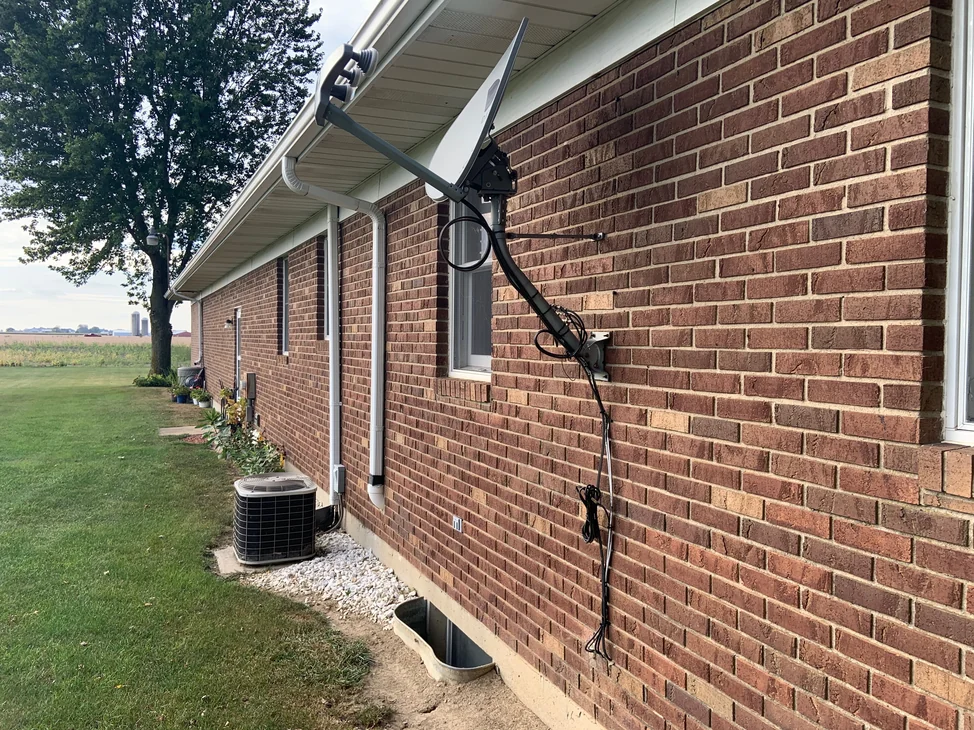 The long side of the single-story brick house in 2019, seen down its length from one end, with a satellite dish on the wall and open farmland beyond.