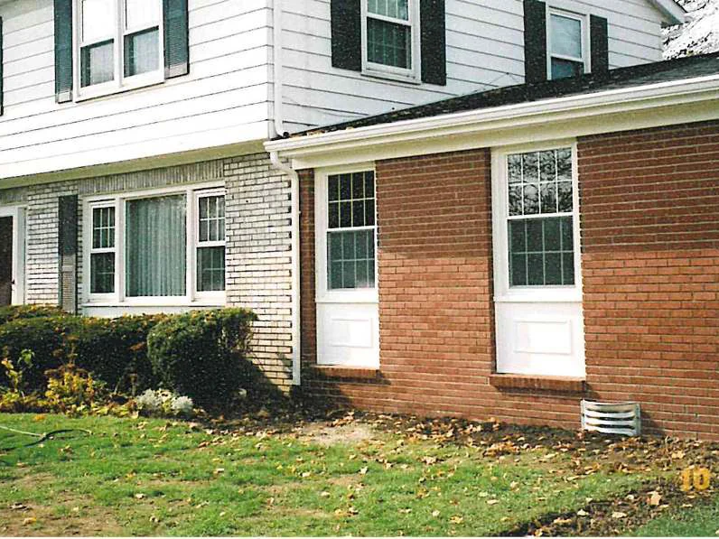 The new addition in 2003, a wall of plain red brick meeting the cream brick of the original house at a flush corner under the same white siding