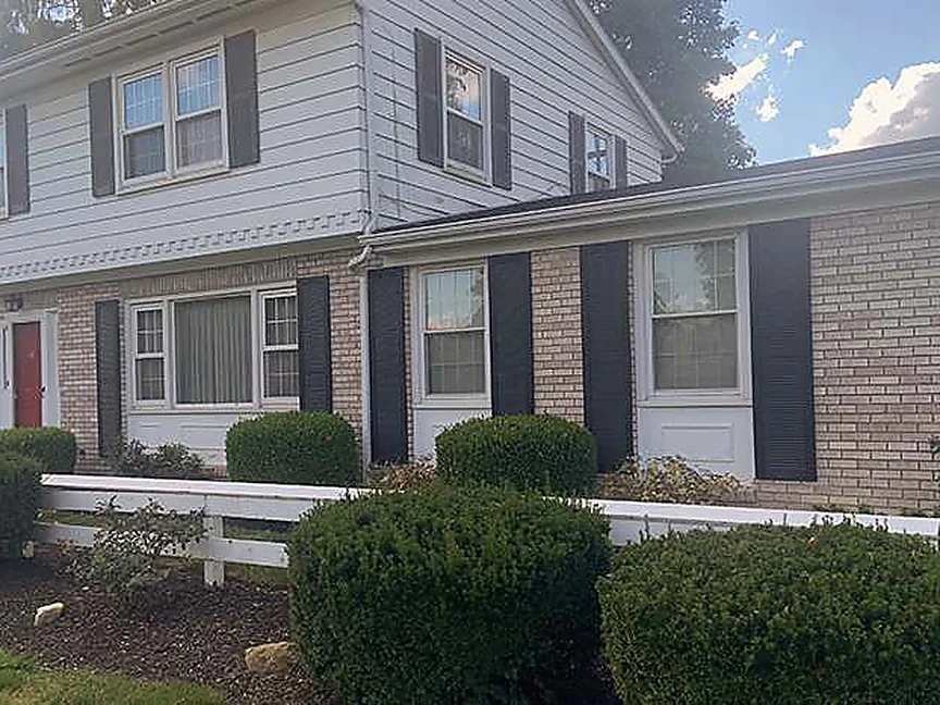 The same elevation closer in 2019, the addition's brick carrying past the bay window into the original wall with a white rail fence and grown-in shrubs along the front