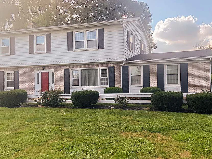 The whole front of the house in 2019, the original brick and the single-story addition on the right reading as one cream range under white siding and dark shutters