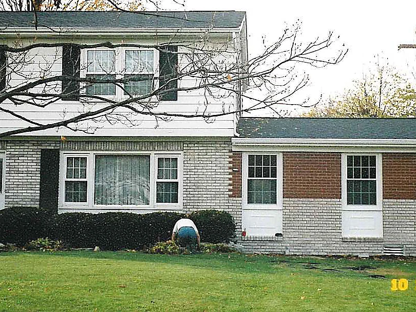 The house partway through the work in 2003, the lower courses of the addition already carried to cream while a band of red brick is still untreated above them, an applicator kneeling at the bed in front