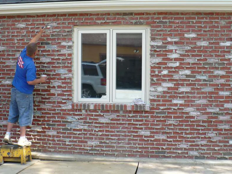 An applicator standing on a step stool in 2008, working stain into the same wall by hand with a small brush and a cup in his other hand