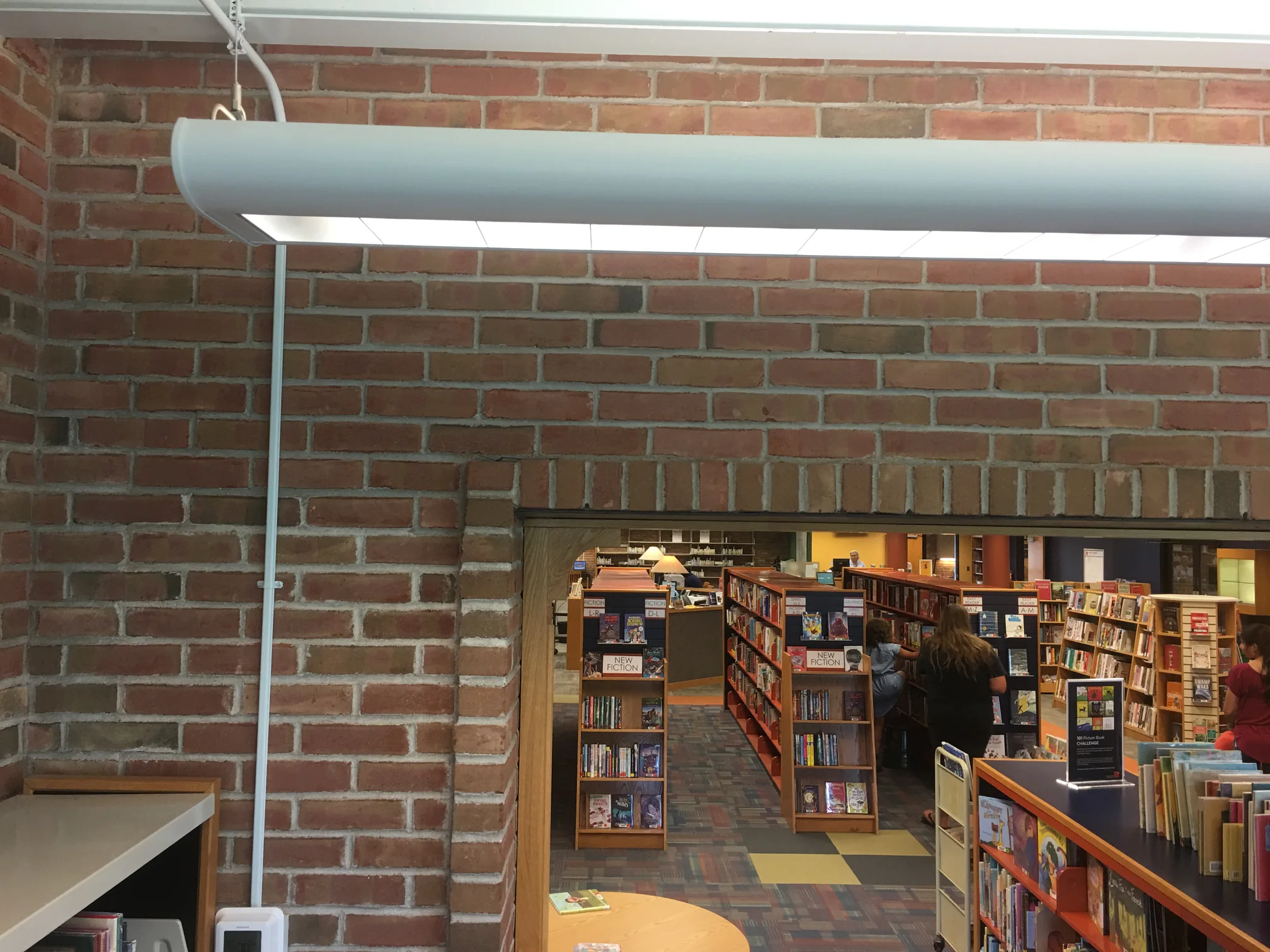 An interior brick wall in the library in 2019, with a wide opening through to the shelves where people are browsing