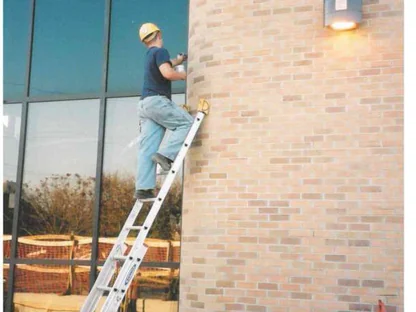 An applicator in a hard hat on an extension ladder against one rounded pier in October 2001, working stain into the brick by hand beside a bay of glass