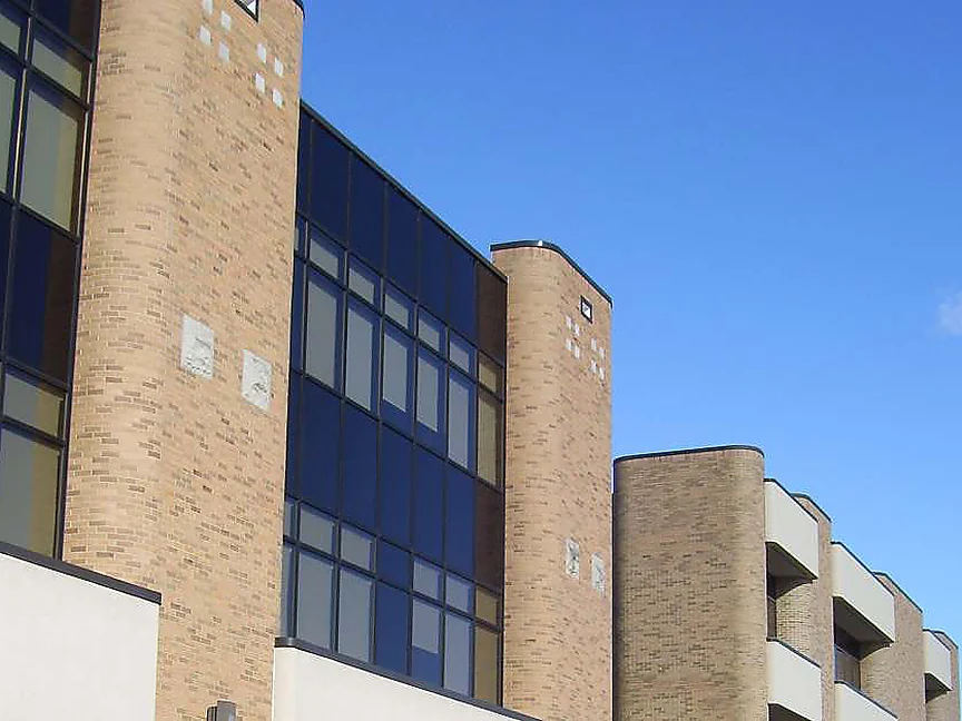 The upper wall of the hospital in 2019, rounded buff brick piers between bays of dark glass under a clear sky, a lower wing of the same brick beyond