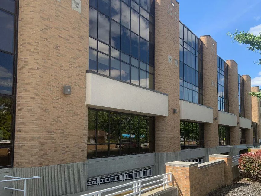 The highway side of the hospital in 2019 from the foot of the entrance ramp, rounded brick piers and glass bays above a concrete base, a white railing and a low brick planter in front