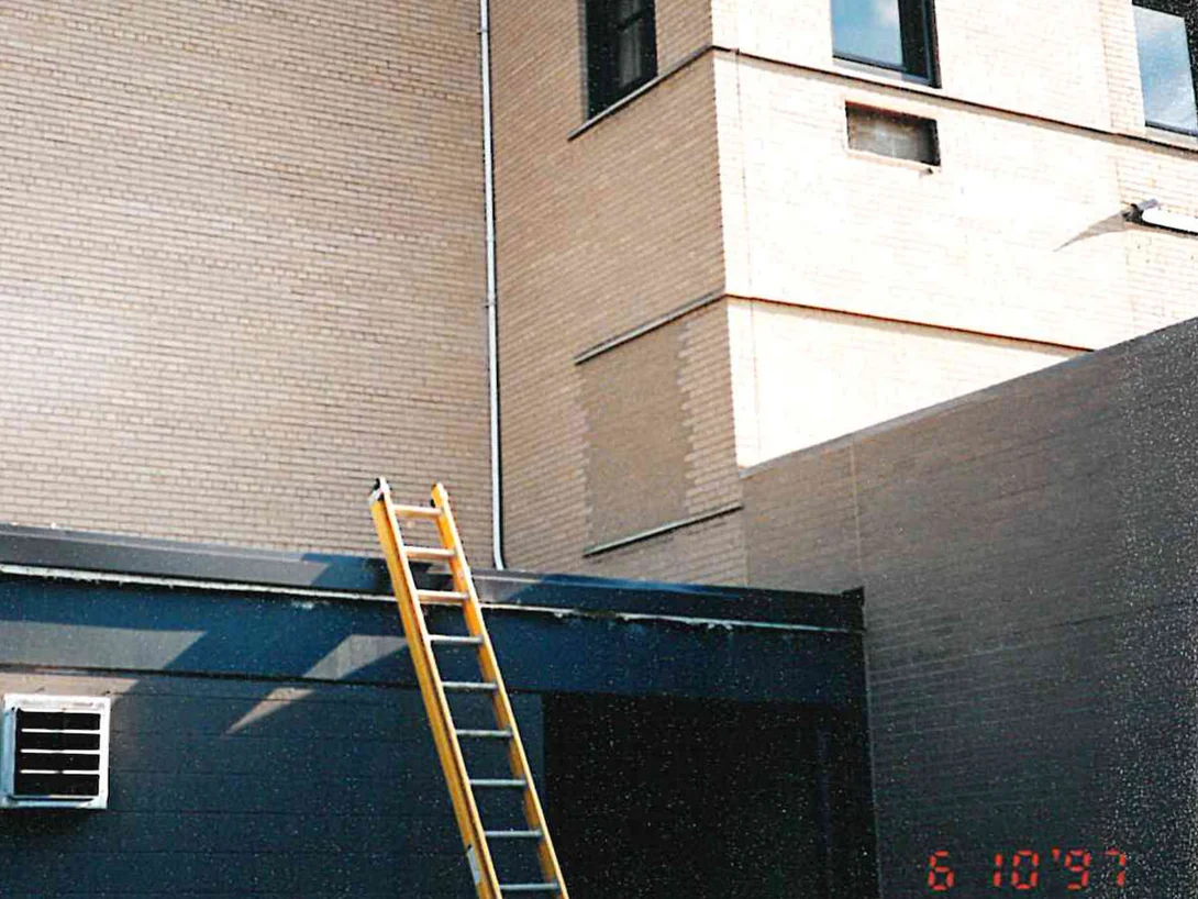 A tall blank wall of brick on the medical center in 1997, its color reading grey beside the warmer cream brick of the corner block next to it, with an extension ladder up to the roof below