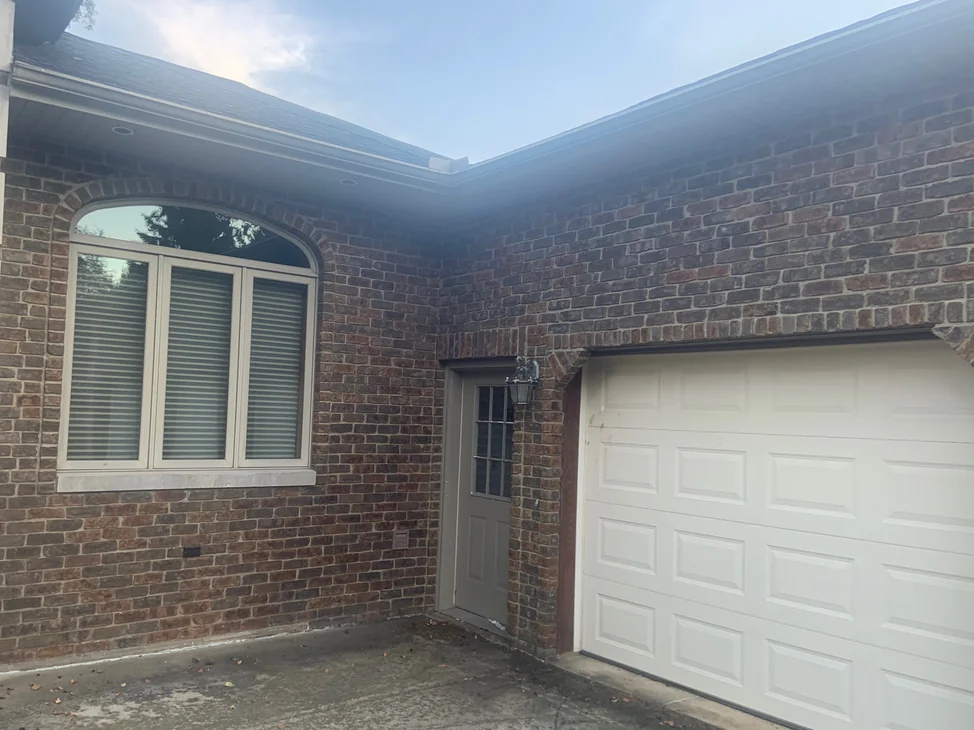 The outside corner of the house in 2019, an arched triple window on one face and a service door and garage door on the other, the brick an even red and brown range across both.