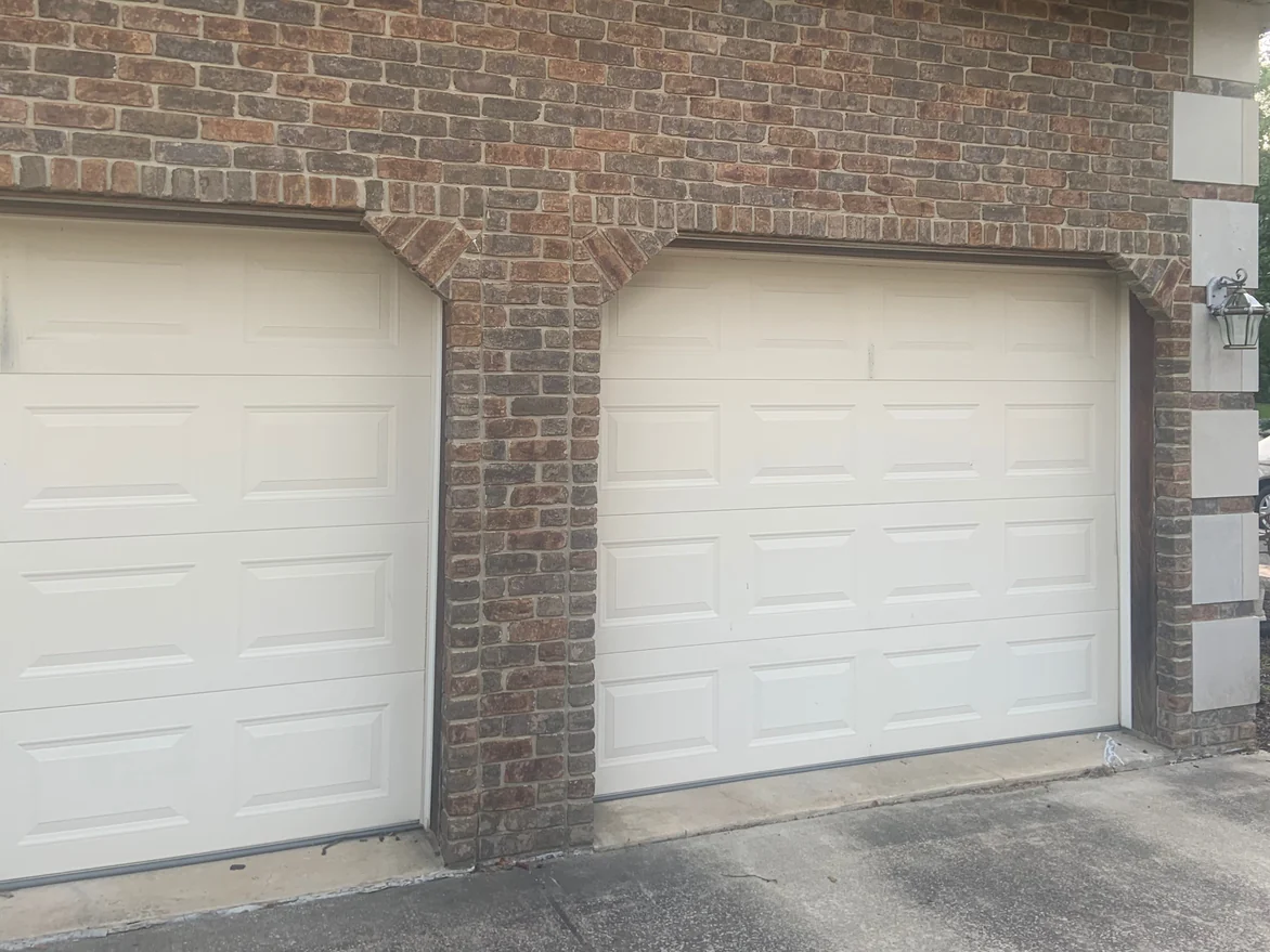 Two white garage doors set in blended red and brown brick under a soldier course, with a cast stone pier and a carriage lamp at the corner