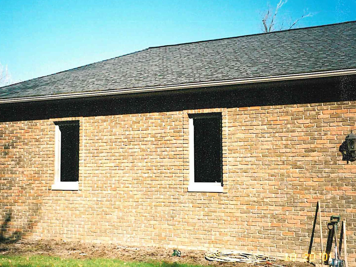 A long single-story elevation in pale tan brick with two tall narrow windows, garden tools leaning against the wall at the right