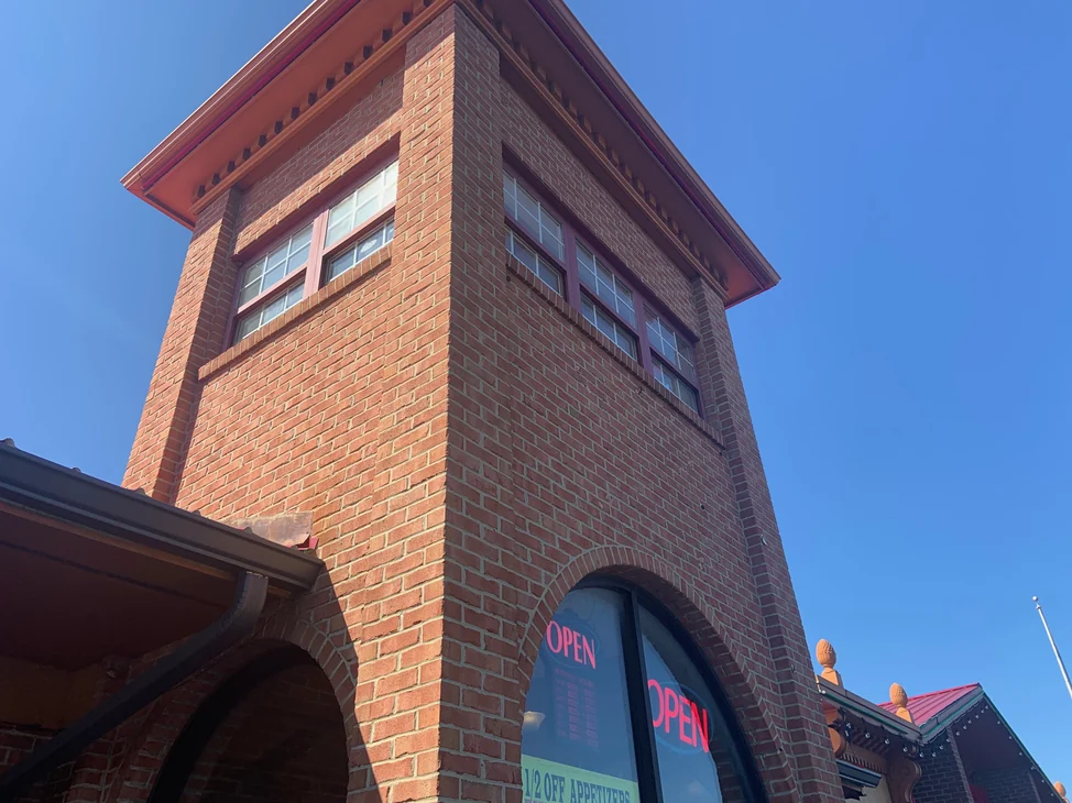 The entry tower from the pavement in 2019, two faces of red brick meeting at the corner under a projecting cornice against a clear sky