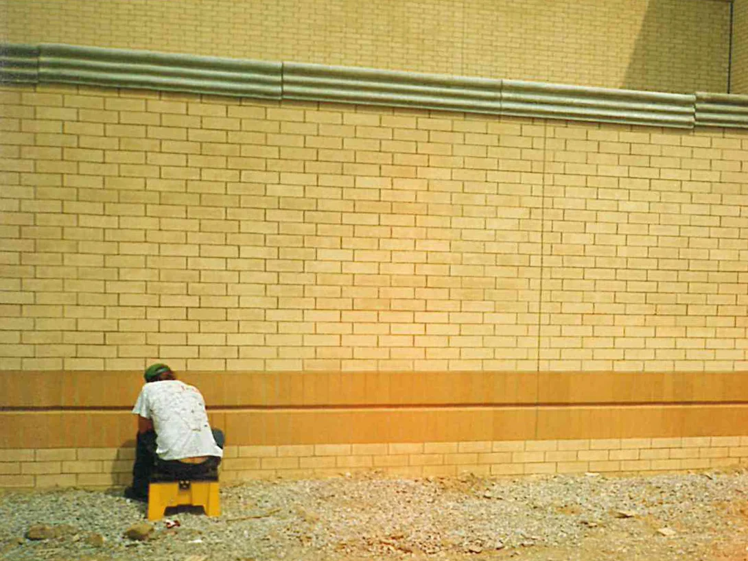 An applicator seated on a step stool in 1996 working along the base soldier band of a low buff brick wall, the ground still unfinished