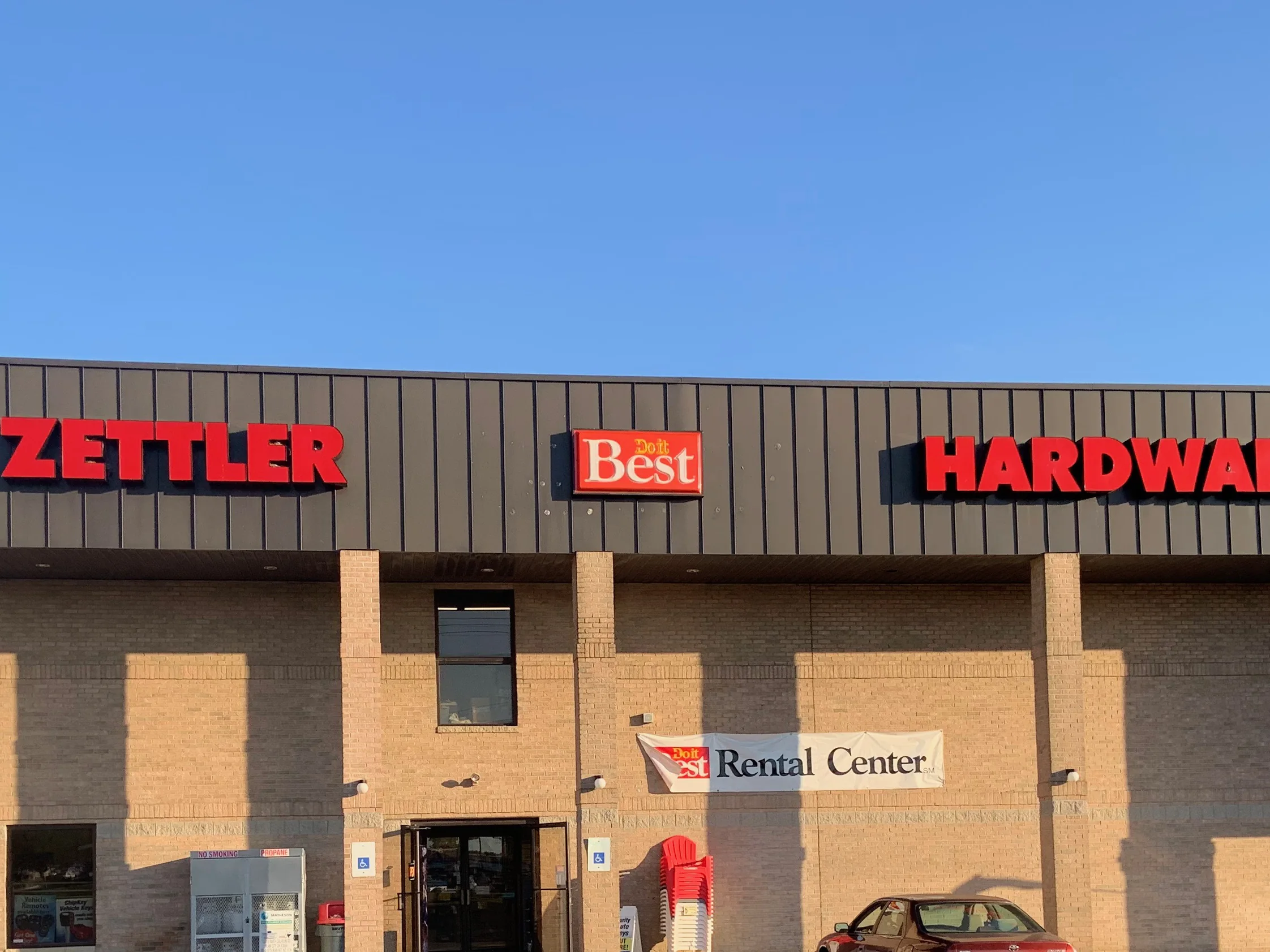 The hardware store seen across its parking lot in 2019, tan brick piers and field walls carrying a dark metal fascia above the entrance