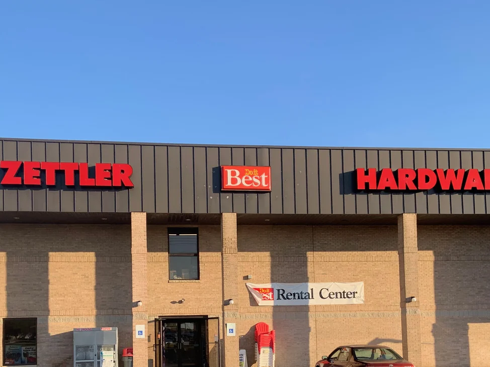The hardware store seen across its parking lot in 2019, tan brick piers and field walls carrying a dark metal fascia above the entrance.