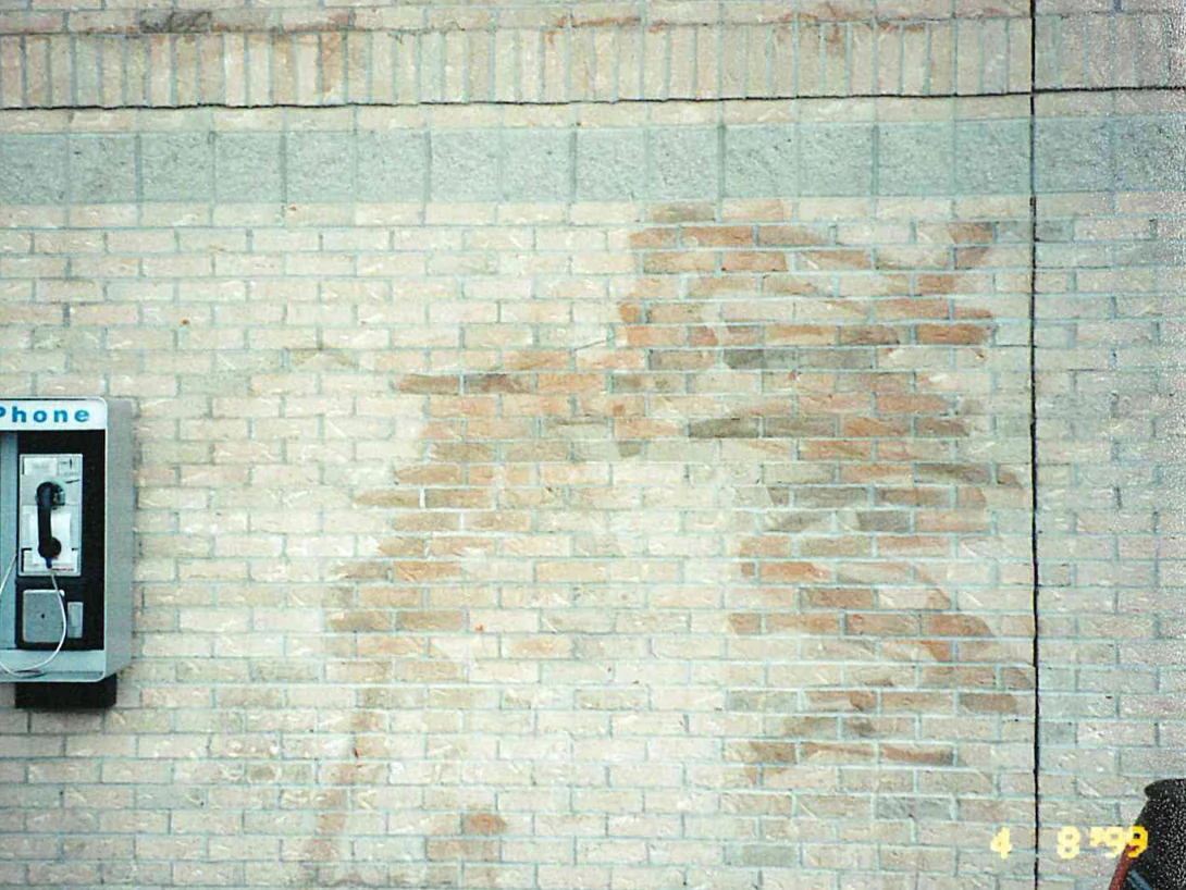 A buff brick wall in 1999 with broad tan and orange patches where a pressure washer has taken the color off, a payphone mounted at the left