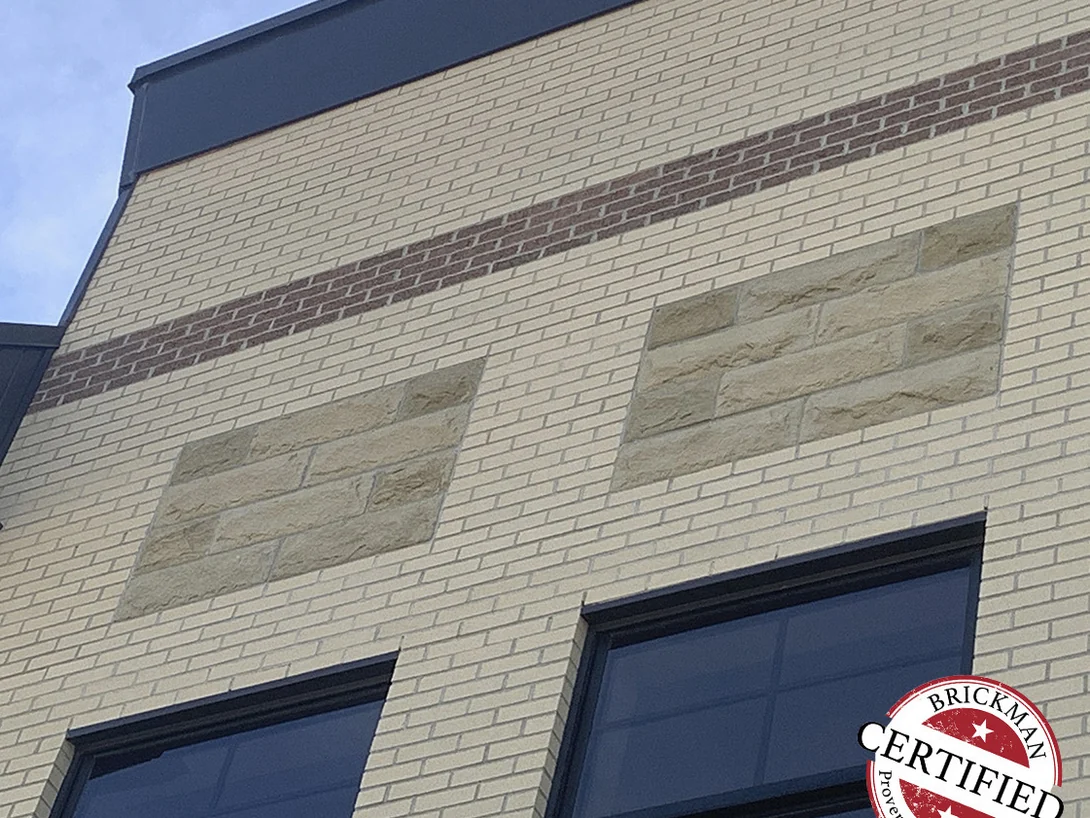 The upper wall in 2019, a red brown accent course running above two panels of cast stone set into the cream brick, with the certification stamp in the corner