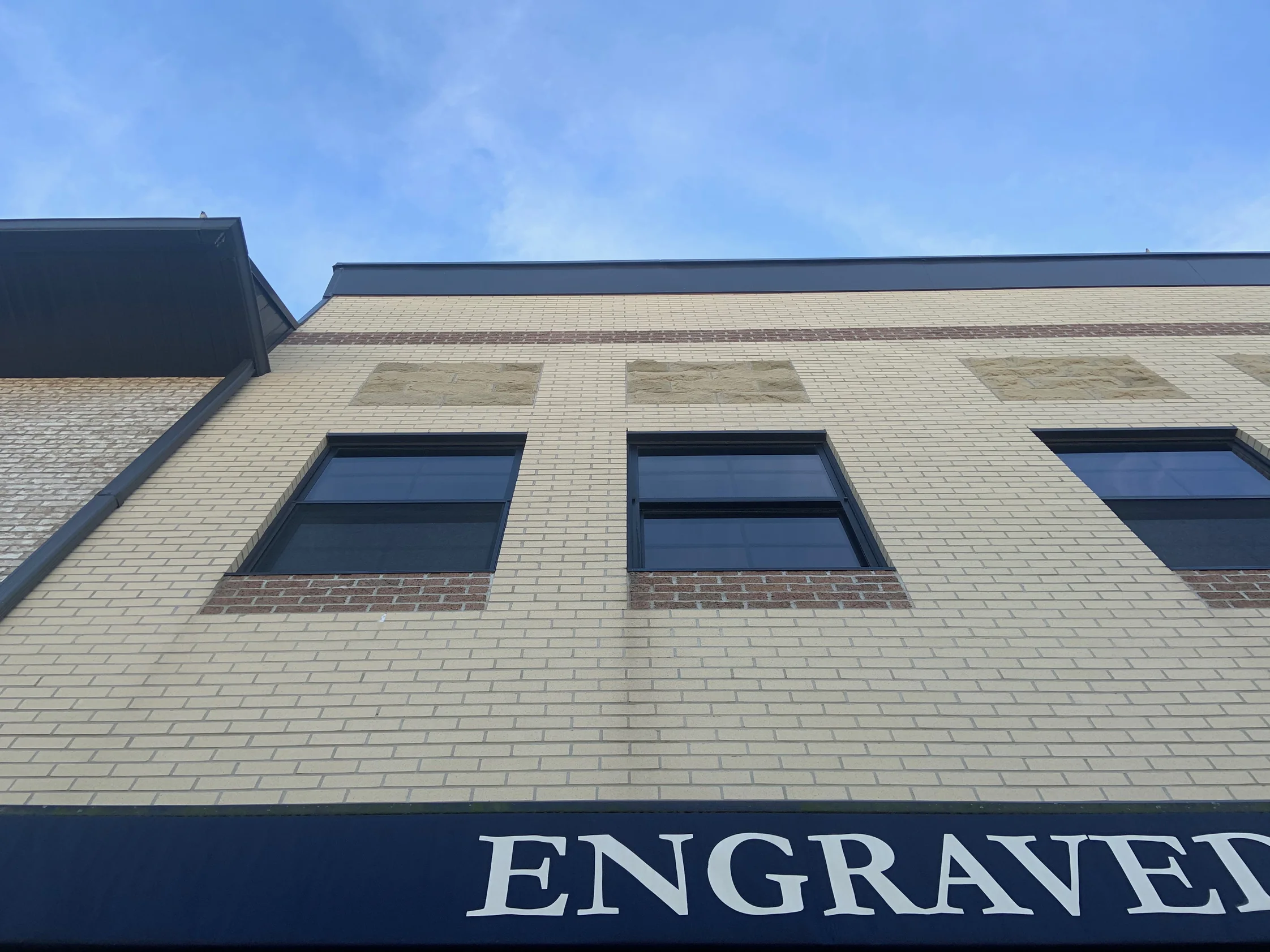 Two windows in 2019 with red brown brick sills and cast stone panels above them, over a dark awning carrying part of a tenant sign