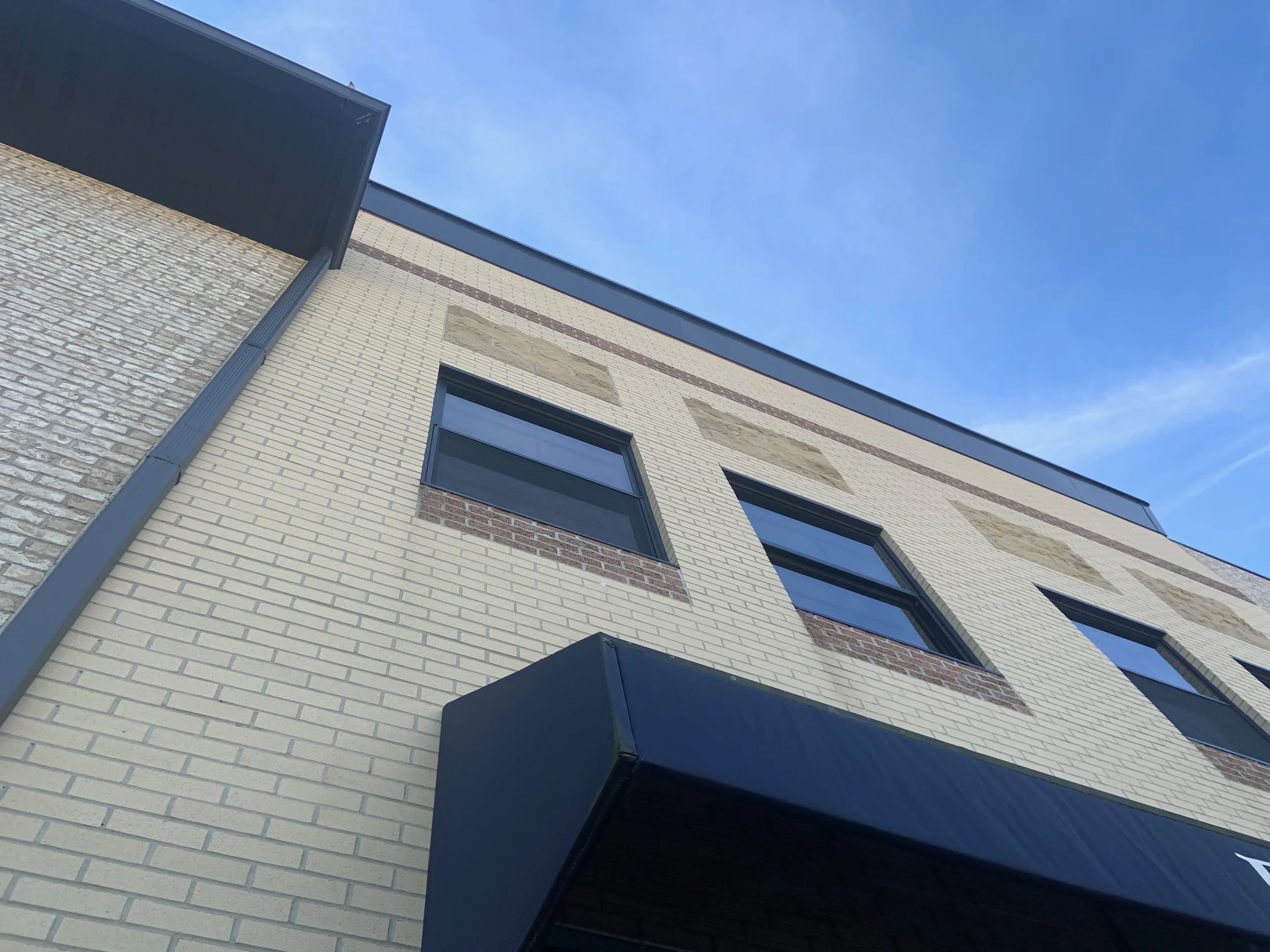 The outside corner of the two-story building in 2019, cream brick running past three window bays to a dark metal parapet with an awning below