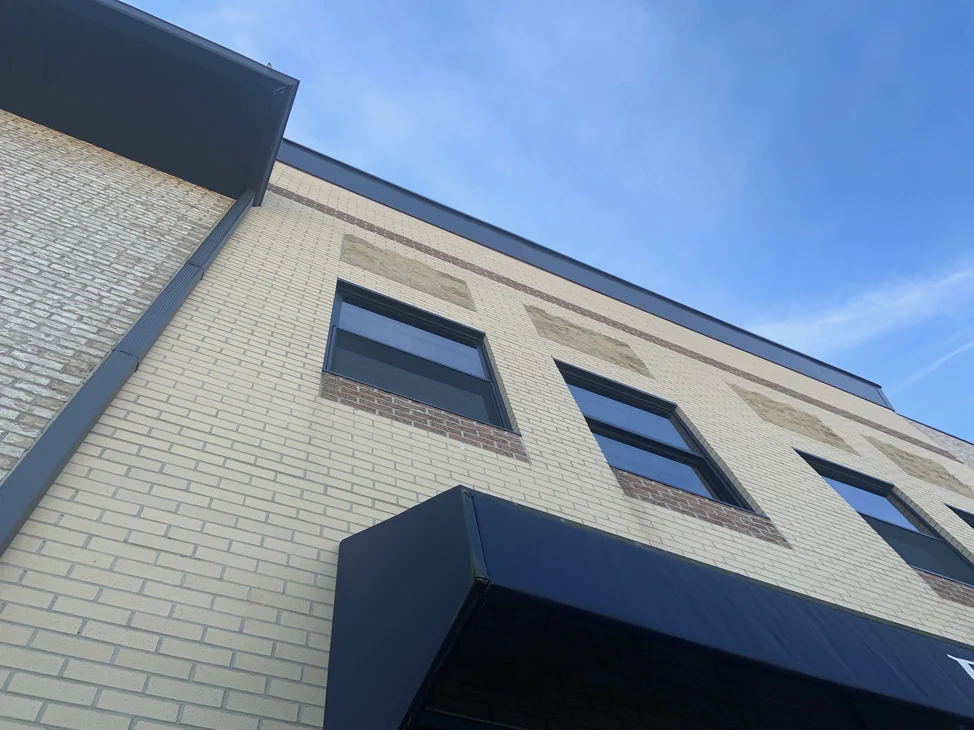 The outside corner of the two-story building in 2019, cream brick running past three window bays to a dark metal parapet with an awning below.
