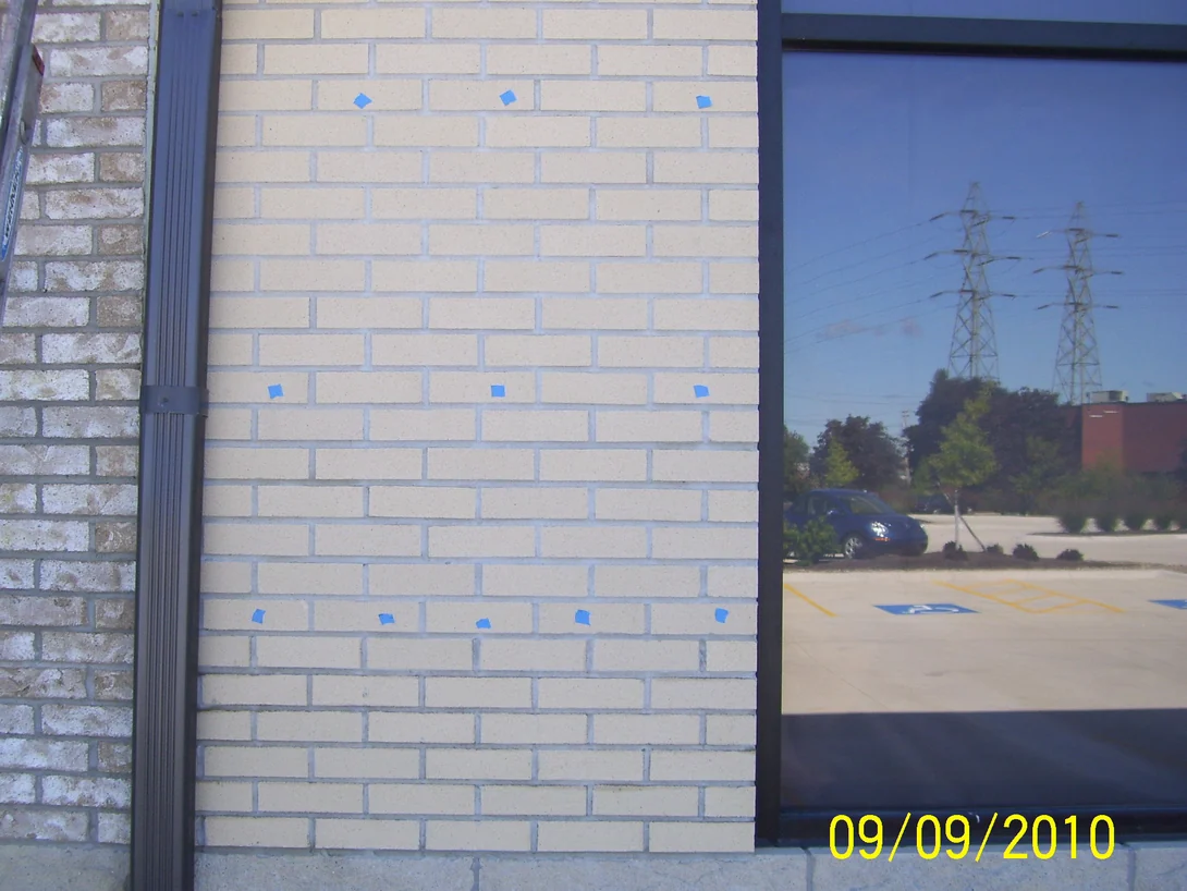 A pier of pale cream brick beside a rougher grey and tan brick wall in 2010, with blue tape squares marking units across the face