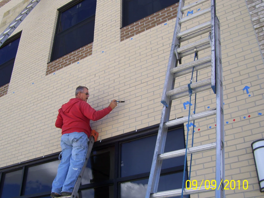 An applicator on a ladder working stain into the cream brick with a hand brush, extension ladders and rope rigged across the wall beside him