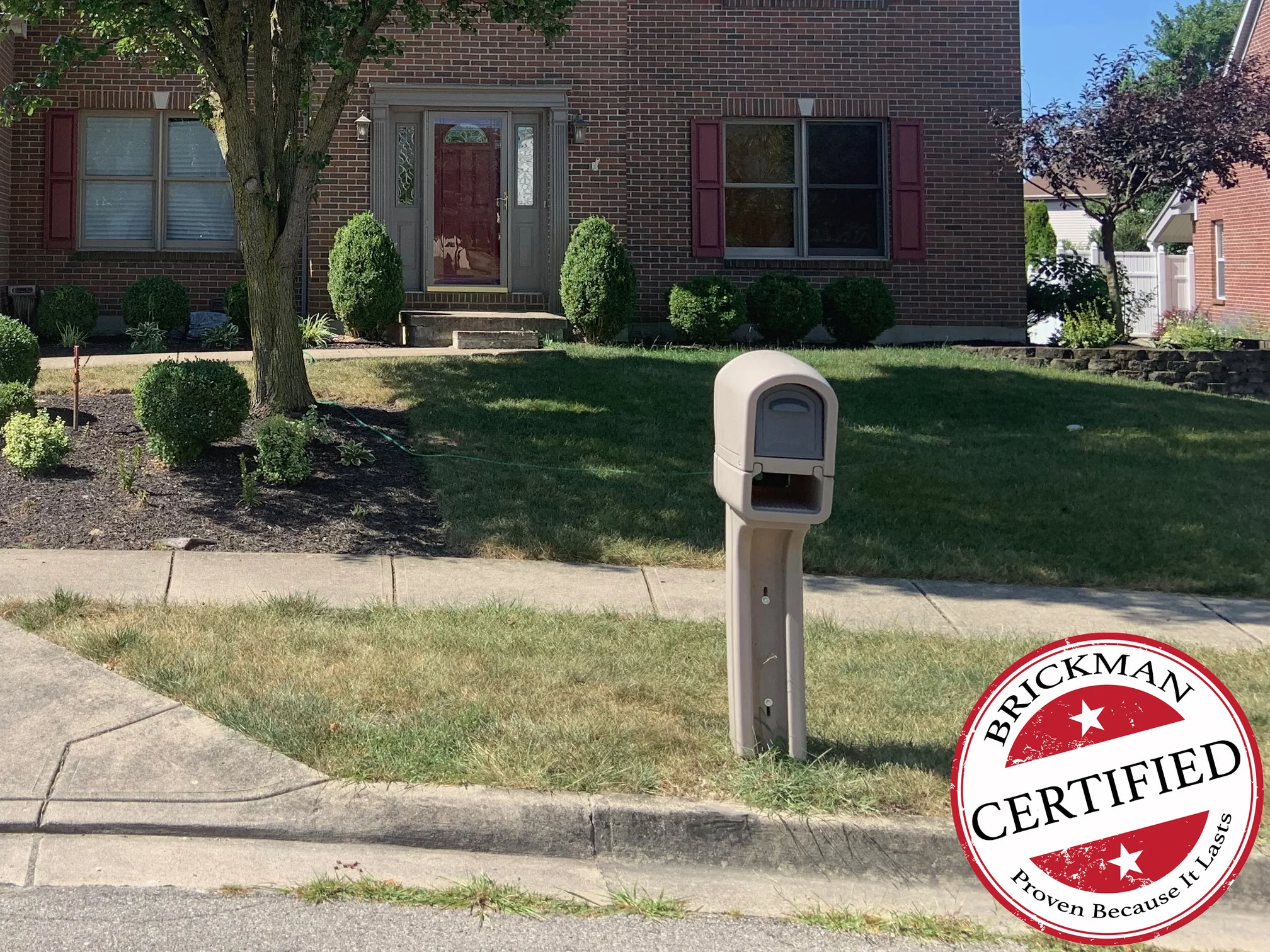 The front entry and the two-story wing beside it in 2019, a grown tree on the lawn and the brick behind it reading as an even range of reds and browns from the ground to the eaves, with the certification stamp at the corner of the frame