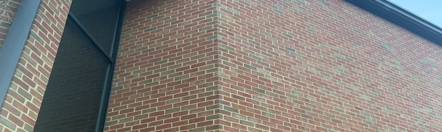 An outside corner of the university building in 2019, red brick with scattered darker units carrying evenly around the corner to a dark roof edge against the sky