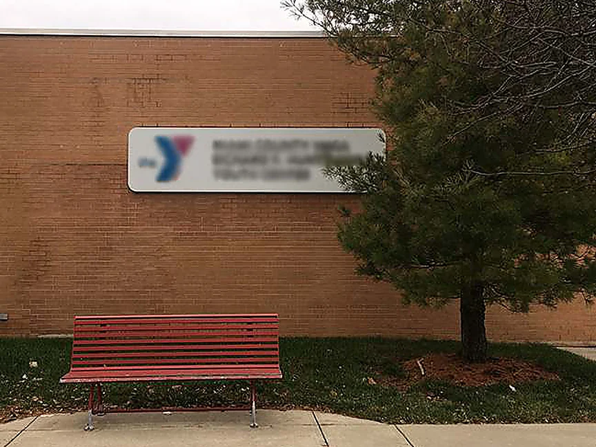 The same wall in 2019 from the walk, brick reading as one range behind the building sign, the red bench still in place and the sapling beside it grown into a pine