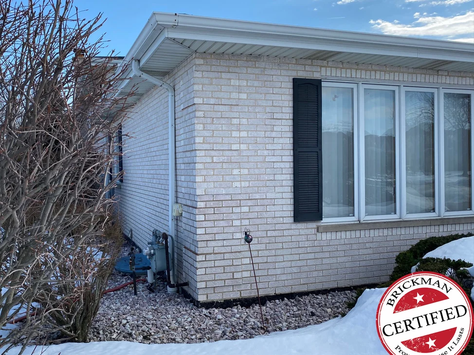 The outside corner of the single-story wing in 2021, pale brick with one light mortar color across both walls, a black shutter beside a bank of windows and the certification stamp in the corner.