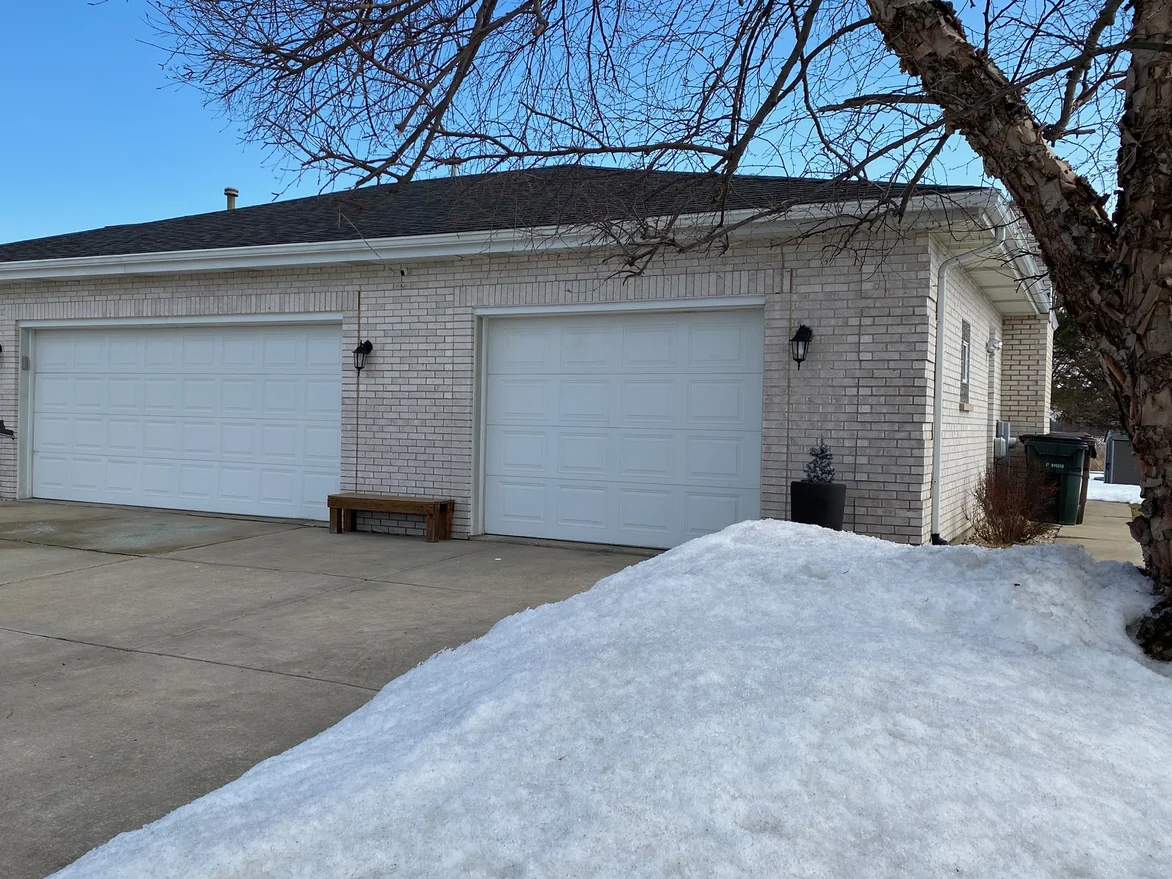 The garage elevation in 2021, two white doors set in pale brick under a low hip roof, a wooden bench between them and a bank of ploughed snow across the driveway