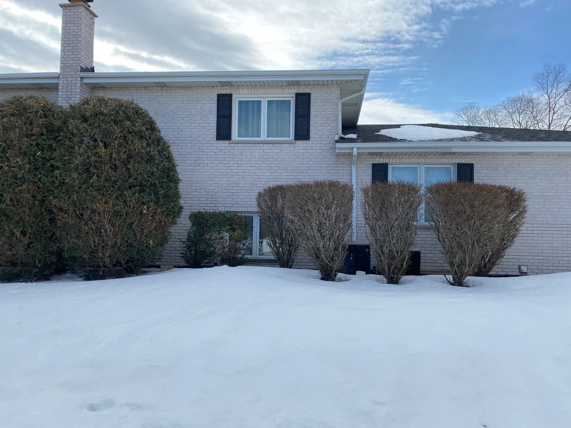 The two-story section of the house in 2021, pale brick with a shuttered upper window and a chimney at the left, the single-story wing stepping down at the right, bare shrubs and deep snow in front